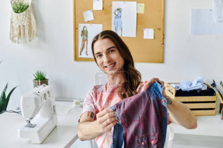 Téléchargez les photos : Un jeune homme sourit en montrant une chemise restaurée dans son atelier de restauration de vêtements de bricolage. - en image libre de droit