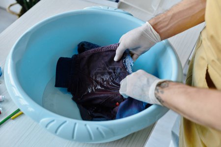 Ein junger Mann mit Handschuhen wäscht ein ausrangiertes Kleidungsstück in einem blauen Becken. Er arbeitet in seinem Atelier, wo er alter Kleidung neues Leben einhaucht.