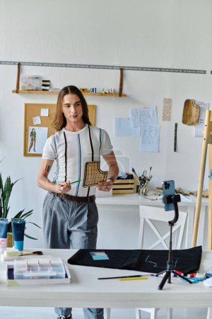 Un jeune homme, vêtu d'une chemise rayée et d'un pantalon gris, se tient dans son atelier, prêt à créer une déclaration de mode durable.