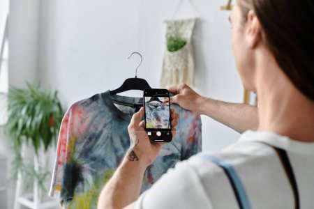 A young man takes a photo of a brightly colored tie-dye shirt he has restored in his clothing atelier.