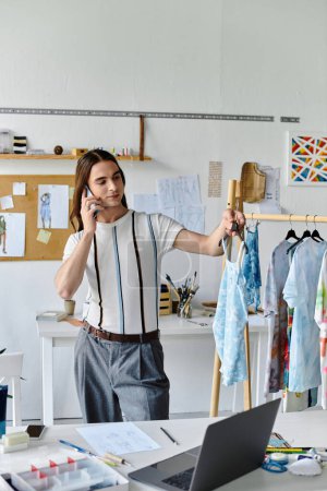Un jeune homme gay dans un atelier de restauration de vêtements de bricolage, au téléphone, présentant un vêtement hes réorienté pour une ligne de vêtements durables.