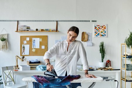 Ein junger Mann im weißen Hemd arbeitet in seinem Atelier an einem Kleiderrestaurierungsprojekt, indem er einem ausrangierten Kleidungsstück mit einem Bügeleisen neues Leben einhaucht.