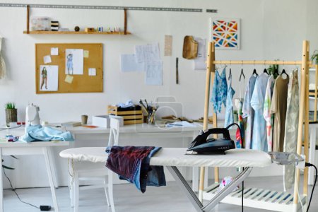 Un atelier à parois blanches avec une machine à coudre, une planche à repasser et un porte-vêtements.