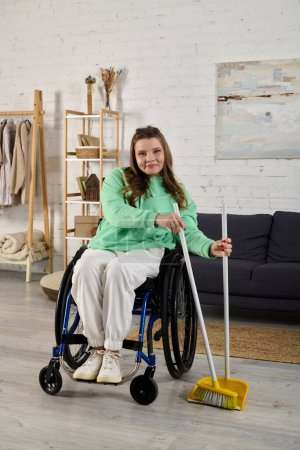 Eine junge Frau im Rollstuhl lächelt, als sie einen Besen in ihrem Wohnzimmer hält.