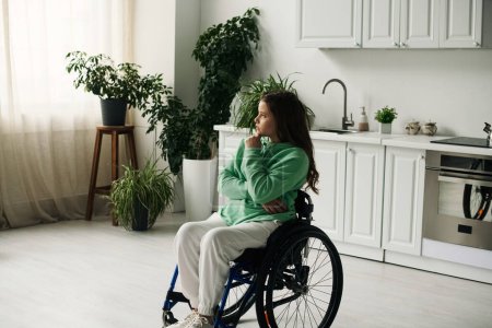 Eine junge Frau im Rollstuhl sitzt gedankenverloren in ihrem Wohnzimmer.