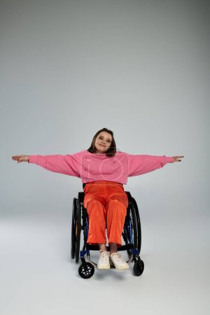 Eine junge brünette Frau in rosa Pullover und orangefarbener Hose sitzt im Rollstuhl, die Arme ausgestreckt, vor einem schlichten grauen Hintergrund..