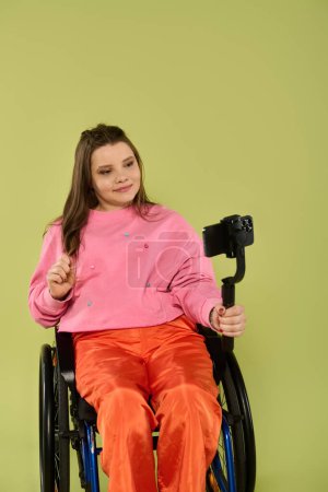A young woman with long brown hair sits in a wheelchair in a studio setting, wearing casual attire and a bright smile.