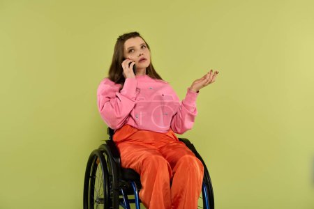 A young woman with long brown hair sits in a wheelchair in a studio setting, talking on the phone while gesturing with her free hand.
