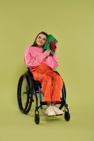 Une jeune femme brune assise dans un fauteuil roulant tient un cadeau enveloppé dans du papier vert avec un ruban rouge. Elle sourit et regarde la caméra..