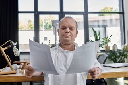 Un homme avec inclusivité est assis à un bureau dans un bureau bien éclairé, examinant attentivement les documents.