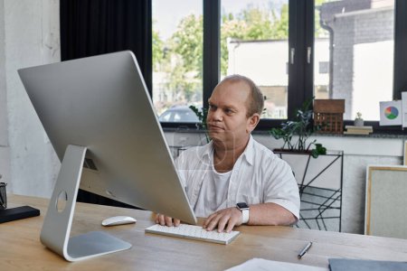 A man with inclusivity sits at a desk in a modern office, working on a computer.