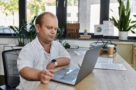 A man with inclusivity sits at his desk in an office, working on his laptop.