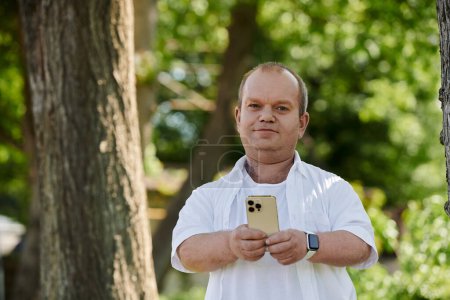 A man with inclusivity with a smartphone in his hand walks through a lush green park on a sunny day.