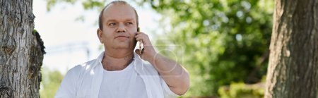 Ein Mann mit weißem Hemd steht an einem Baum und schaut nachdenklich auf sein Handy..