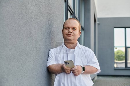 Un homme inclusif dans une chemise blanche se tient dans une ville moderne, tenant un smartphone tout en s'appuyant contre un mur.