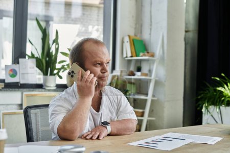 Ein Mann mit weißem Hemd sitzt an einem Schreibtisch und telefoniert in einem hell erleuchteten Büro.