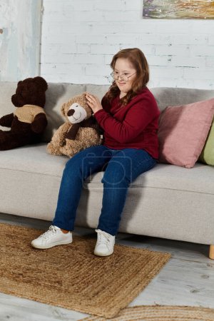 A little girl with Down syndrome sits on a couch with two teddy bears.