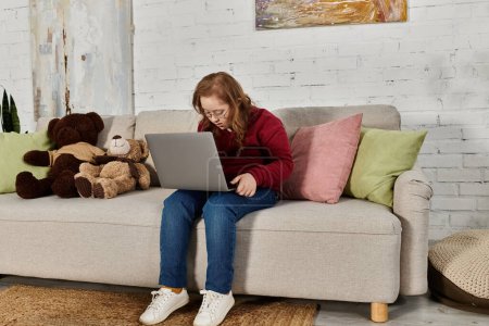 A little girl with Down syndrome sits on a couch at home, using a laptop.