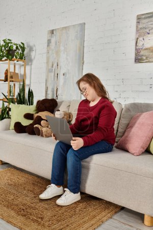 A little girl with Down syndrome sits on a couch at home using a laptop.