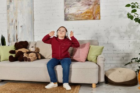 A little girl with Down syndrome sits on a couch at home, wearing headphones and dancing to music.