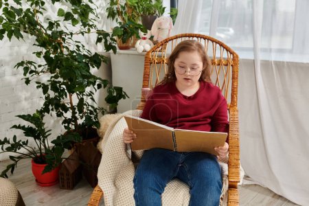 A little girl with Down syndrome sits in a wicker chair, engrossed in a book.