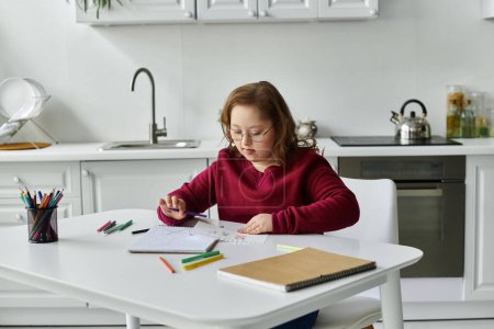A little girl with Down syndrome sits at a kitchen table, drawing in a notebook.