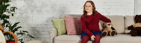 A little girl with Down syndrome sits on a couch at home.