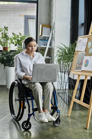 A young businesswoman in a wheelchair works on her laptop in a modern office setting.