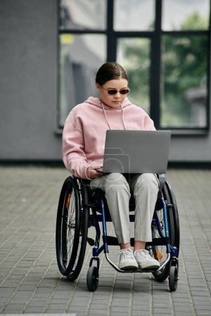 A young woman wearing sunglasses and a pink hoodie sits in a wheelchair outside while using a laptop.