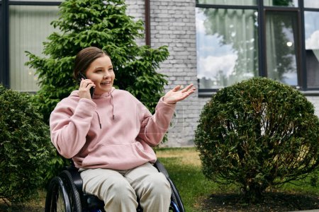 Una joven en una sudadera rosa se sienta en una silla de ruedas afuera, hablando por teléfono y sonriendo.