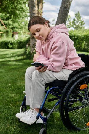 Une jeune femme est assise dans un fauteuil roulant dans un parc herbeux, regardant son téléphone.