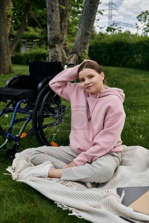 Eine junge Frau in rosa Kapuzenpulli sitzt im Rollstuhl in einem Park, umgeben von viel Grün.