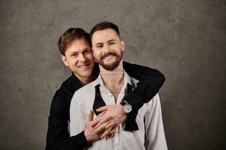 Two men in suits embrace and smile against a grey backdrop.