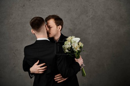 Zwei Männer in eleganten Anzügen stehen dicht beieinander, einer hält einen Strauß weißer Blumen in der Hand..