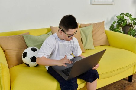 A young boy with Down syndrome sits on a yellow couch using a laptop.