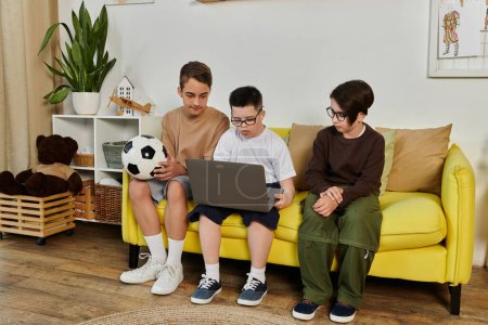 Three young boys sit on a yellow sofa, one looking at a laptop while the other two watch.