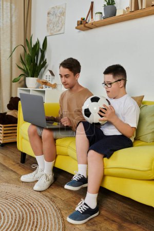Zwei Jungen, einer davon mit Down-Syndrom, sitzen auf einer gelben Couch und schauen am Laptop Fußball.