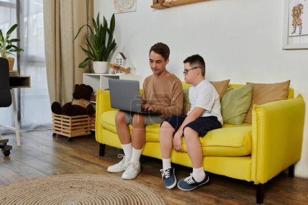 Two boys, one with Down syndrome, are sitting on a yellow sofa at home, using a laptop.