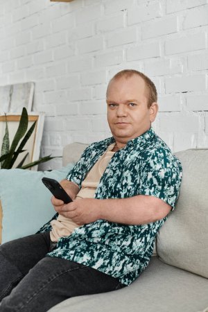 A man with inclusivity sits on a couch, holding a phone and looking intently at the screen.