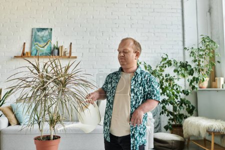 A man with inclusivity in a green shirt waters a large houseplant in a white room.