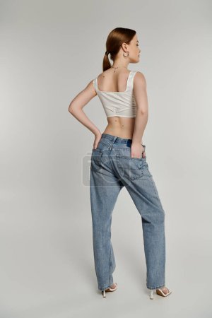 A young woman with her back to the camera poses in light-wash denim jeans and a white crop top.