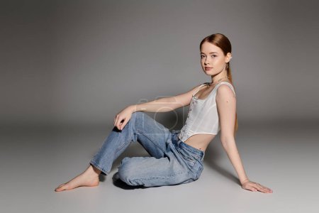 A young woman sits on a gray backdrop, wearing a white corset and denim jeans.