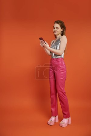 A young woman in a silver top and pink pants looks at her phone in front of an orange backdrop.