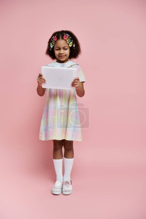 A young African American girl in a colorful dress stands on a pink background and looks intently at a tablet.