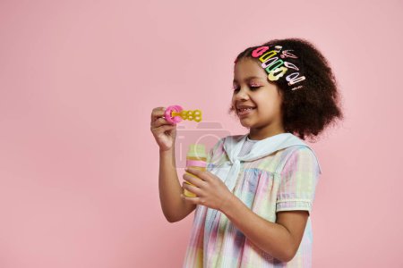 A young African American girl in a colorful dress blows bubbles with a pink wand against a pink background.
