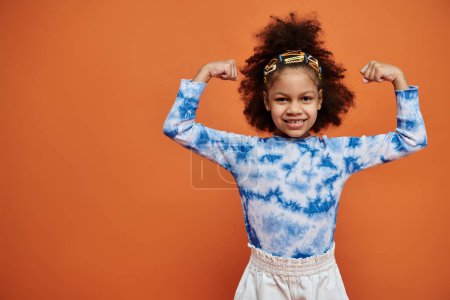 Ein junges afroamerikanisches Mädchen mit stylischen Haarspangen und Krawattenfärberhemd lässt vor orangefarbenem Hintergrund ihre Muskeln spielen.