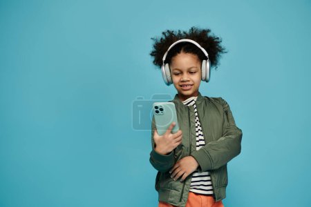 A young African American girl with curly hair smiles as she looks at her smartphone. She is wearing headphones and a green jacket.