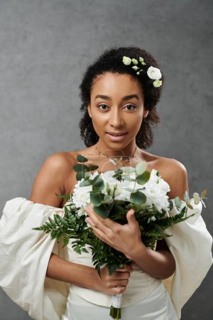 A beautiful African American bride in a white wedding dress and floral hairpiece holds a bouquet of white flowers.