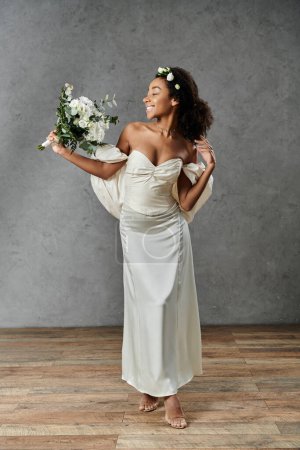 Stunning African American bride in white gown, flowers in hair, smiling, holding bouquet, against grey background.