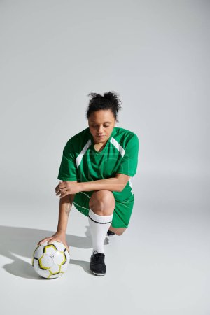 A female athlete in a green jersey kneels with a soccer ball in front of her.